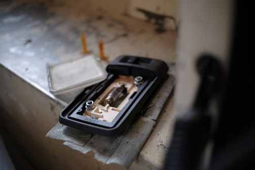 A close-up view of a disassembled electronic device, possibly a battery compartment, placed on a textured and worn surface. Two cylindrical objects stand upright in the background, adding to the industrial feel.
