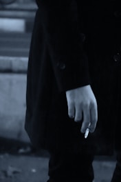 A close-up of a person's hand holding a cigarette. The person is wearing a dark coat, and the image has a muted, monochrome color palette.