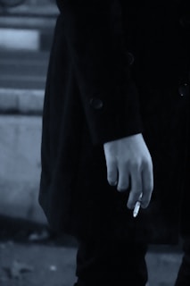 A close-up of a person's hand holding a cigarette. The person is wearing a dark coat, and the image has a muted, monochrome color palette.