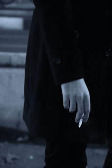 A close-up of a person's hand holding a cigarette. The person is wearing a dark coat, and the image has a muted, monochrome color palette.