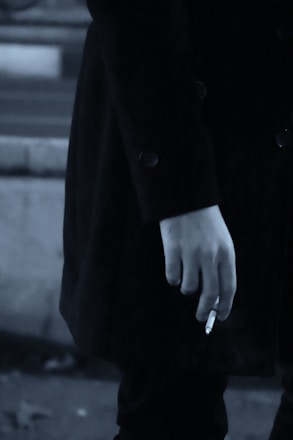 A close-up of a person's hand holding a cigarette. The person is wearing a dark coat, and the image has a muted, monochrome color palette.