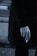 A close-up of a person's hand holding a cigarette. The person is wearing a dark coat, and the image has a muted, monochrome color palette.