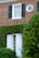 A brick house facade with a rectangular window featuring black shutters and a circular window above. Below the windows, a white door with glass panes is partially covered by green ivy and surrounded by well-maintained bushes and foliage.