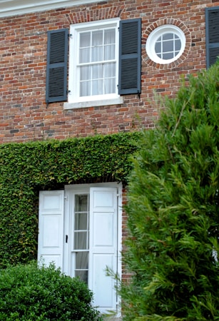 A brick house facade with a rectangular window featuring black shutters and a circular window above. Below the windows, a white door with glass panes is partially covered by green ivy and surrounded by well-maintained bushes and foliage.