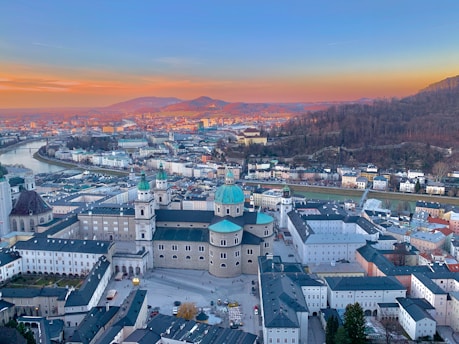An aerial view of a historic European cityscape at sunset, featuring classical architecture with green-topped domes and river winding through the urban landscape. The city is nestled between gently rolling hills, with autumnal colors visible on the nearby hills.