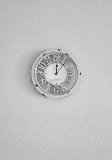 Artisanal wall clock with a simple round face and subtle warm hues, hanging on a textured light gray wall.
