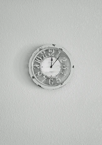 Artisanal wall clock with a simple round face and subtle warm hues, hanging on a textured light gray wall.