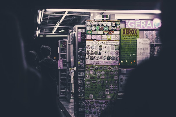 A dimly lit street kiosk displays a variety of vibrant stickers and posters on the front panel. The interior is illuminated by fluorescent lights, casting a moody glow. Two figures stand in front of the kiosk, partially silhouetted against the light.