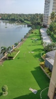 A pristine landscaped area featuring a neatly mowed green lawn bordered by a row of ornamental palm trees. Adjacent to the lawn is a body of water, possibly a lake or river, with dense greenery in the background. A tall residential building is visible on the right, and a small playground with a swing set is located on the lawn. The scene is lit with bright daylight, showcasing a clear sky.