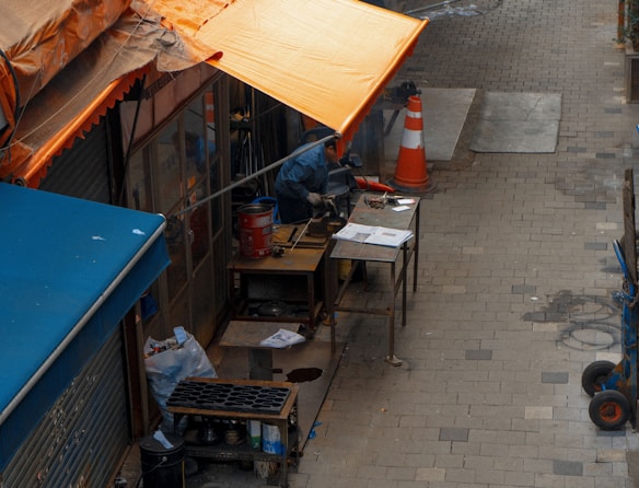 An outdoor workspace is situated along a paved alley, partially covered by an orange canopy. A person is engaged in working with machinery on a table, with tools and papers scattered around. The scene includes a traffic cone and multiple tables, one of which holds a red container.