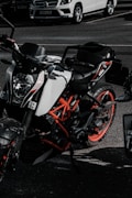A sporty, black and white KTM motorcycle with orange accents is parked in a lot. Its sleek, angular design is highlighted by the dark and moody lighting. A white Mercedes-Benz SUV is visible in the background, partially obscured, parked alongside the motorcycle.