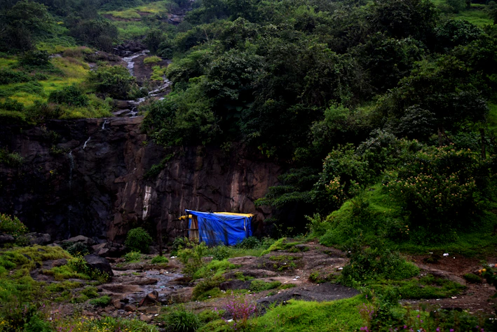 A sleek green shower tent pitched beside a flowing mountain stream under a bright blue sky.