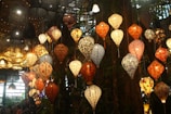An array of colorful lanterns hangs from the ceiling, creating a vibrant and warm ambiance. Each lantern is uniquely designed with intricate patterns in various colors such as red, blue, and white. The background features a lush, natural setting with trees and foliage, adding a touch of nature to the decorative display.