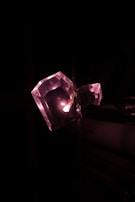 A faceted, heart-shaped crystal is illuminated from within by soft pink light against a dark background. The lighting creates a shimmering effect on the crystal's surface, highlighting its geometric facets.