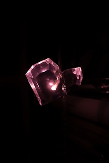 A faceted, heart-shaped crystal is illuminated from within by soft pink light against a dark background. The lighting creates a shimmering effect on the crystal's surface, highlighting its geometric facets.