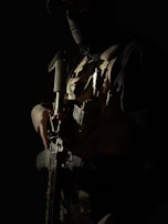 A person wearing tactical gear and a mask stands in a dimly lit environment. They are holding a firearm, and the lighting creates dramatic shadows across their face and body. The details of the tactical vest, including various pouches and equipment, are partially visible.