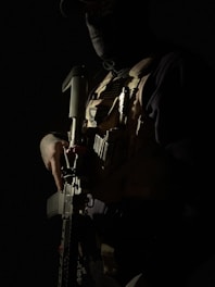 A person wearing tactical gear and a mask stands in a dimly lit environment. They are holding a firearm, and the lighting creates dramatic shadows across their face and body. The details of the tactical vest, including various pouches and equipment, are partially visible.