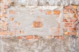 An old brick wall with patches of plaster covering various areas. The wall has a weathered and aged appearance, with some bricks partially exposed and others fully covered by crumbling plaster. The bricks are of varying shades of red and orange, while the plaster is a dull gray. The surface shows signs of deterioration, with chips and holes visible throughout.