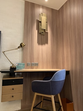 A modern workspace with a wooden desk and a blue upholstered chair. A wall-mounted decorative piece adds an artistic touch above the desk. The wall is paneled with wood, contributing to a warm and inviting atmosphere. A brass desk lamp sits to the side, and a small cabinet with drawers is attached under the desk. Electrical outlets are visible on the wall, and a waste bin is at the corner.