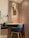 A modern workspace with a wooden desk and a blue upholstered chair. A wall-mounted decorative piece adds an artistic touch above the desk. The wall is paneled with wood, contributing to a warm and inviting atmosphere. A brass desk lamp sits to the side, and a small cabinet with drawers is attached under the desk. Electrical outlets are visible on the wall, and a waste bin is at the corner.