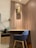 A modern workspace with a wooden desk and a blue upholstered chair. A wall-mounted decorative piece adds an artistic touch above the desk. The wall is paneled with wood, contributing to a warm and inviting atmosphere. A brass desk lamp sits to the side, and a small cabinet with drawers is attached under the desk. Electrical outlets are visible on the wall, and a waste bin is at the corner.
