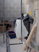 A partially constructed or renovated room with exposed brick walls and unfinished surfaces. A washing machine is positioned against the wall, partially covered by a black tarp. A blue and orange plastic bin is placed next to the machine. There are wires and pipes scattered across the concrete floor, alongside wooden and metal construction materials.