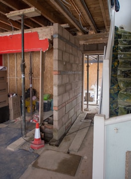 A construction site inside a building with unfinished walls made of concrete blocks. Exposed wires and beams are visible on the ceiling. Various construction materials, including wooden planks, are scattered around. There is a red partition and several cardboard boxes are stacked nearby. A traffic cone is positioned near a support beam.