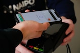 A person is using a smartphone with a mobile payment app to make a contactless payment on a card reader. The phone displays a Mastercard logo and shows the payment screen.