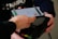 A person is using a smartphone with a mobile payment app to make a contactless payment on a card reader. The phone displays a Mastercard logo and shows the payment screen.