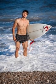 A man stands in the ocean with waves around his legs, holding a surfboard. He is shirtless and wearing black shorts. The surfboard has branded logos and is white with a red design. The background features a vibrant blue ocean.