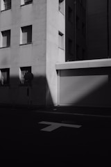 A black and white photograph captures an urban street corner. The sharp shadows and highlights accentuate the geometric shapes of a modern building facade with closed windows. A metal roll-up door and a traffic sign are visible, contributing to the somber atmosphere.
