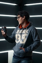 A person wearing glasses and a blue hoodie with the word 'Bronx' and the number '95' printed on it is holding a camera lens in one hand. The background features horizontal light strips that create an illuminated setting. The person has short, curly dark hair and is smiling slightly.