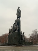 A large monument features a tall statue of a man standing atop a pedestal, surrounded by several smaller statues depicting soldiers and civilians. The scene appears overcast with leafless trees in the background, suggesting a winter or autumn setting.