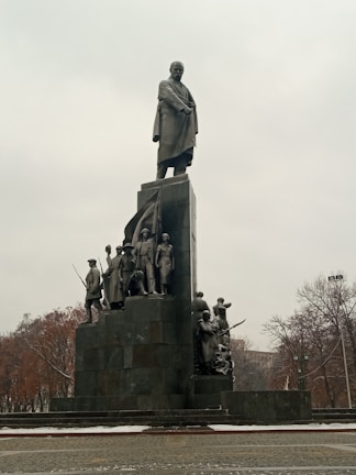 A large monument features a tall statue of a man standing atop a pedestal, surrounded by several smaller statues depicting soldiers and civilians. The scene appears overcast with leafless trees in the background, suggesting a winter or autumn setting.