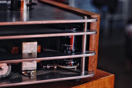 A close-up view of a vintage electronic device, possibly an amplifier, with visible metallic and wooden components. The image highlights the detailed construction of the device, showcasing cylindrical capacitors, metal bars, and wooden panels.