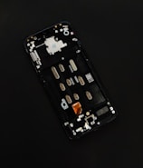 A disassembled smartphone with its internal components visible, showcasing various circuit elements such as metal connectors, screws, and a QR code on a circuit board. The background is solid black, drawing focus to the partially exposed circuitry.