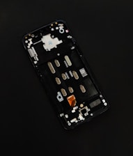 A disassembled smartphone with its internal components visible, showcasing various circuit elements such as metal connectors, screws, and a QR code on a circuit board. The background is solid black, drawing focus to the partially exposed circuitry.