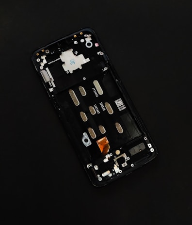 A disassembled smartphone with its internal components visible, showcasing various circuit elements such as metal connectors, screws, and a QR code on a circuit board. The background is solid black, drawing focus to the partially exposed circuitry.