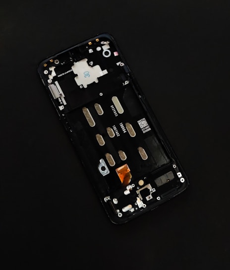 A disassembled smartphone with its internal components visible, showcasing various circuit elements such as metal connectors, screws, and a QR code on a circuit board. The background is solid black, drawing focus to the partially exposed circuitry.
