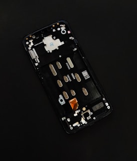 A disassembled smartphone with its internal components visible, showcasing various circuit elements such as metal connectors, screws, and a QR code on a circuit board. The background is solid black, drawing focus to the partially exposed circuitry.