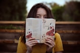 A person with long hair wearing a yellow shirt is holding a book titled 'The Handmaid's Tale' by Margaret Atwood in front of their face. They are sitting outdoors on wooden steps, with a blurred natural background of greenery.