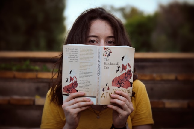 A person with long hair wearing a yellow shirt is holding a book titled 'The Handmaid's Tale' by Margaret Atwood in front of their face. They are sitting outdoors on wooden steps, with a blurred natural background of greenery.