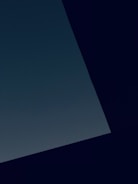 A minimalist composition featuring a large, dark geometric shape, possibly a wall or structure, set against a slightly lighter background. The image creates a stark contrast between the deep, dark colors and the subtle hues of the background.