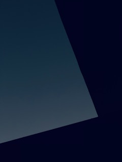 A minimalist composition featuring a large, dark geometric shape, possibly a wall or structure, set against a slightly lighter background. The image creates a stark contrast between the deep, dark colors and the subtle hues of the background.