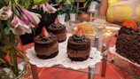 Assorted cakes and desserts are elegantly displayed, with chocolate cakes topped with strawberries on napkins. A larger cake covered in sprinkles is also visible. Fresh flowers and a partially eaten cheesecake add to the festive setup on a red patterned tablecloth.