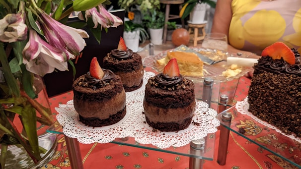 Assorted cakes and desserts are elegantly displayed, with chocolate cakes topped with strawberries on napkins. A larger cake covered in sprinkles is also visible. Fresh flowers and a partially eaten cheesecake add to the festive setup on a red patterned tablecloth.