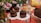 Assorted cakes and desserts are elegantly displayed, with chocolate cakes topped with strawberries on napkins. A larger cake covered in sprinkles is also visible. Fresh flowers and a partially eaten cheesecake add to the festive setup on a red patterned tablecloth.