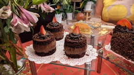 Assorted cakes and desserts are elegantly displayed, with chocolate cakes topped with strawberries on napkins. A larger cake covered in sprinkles is also visible. Fresh flowers and a partially eaten cheesecake add to the festive setup on a red patterned tablecloth.