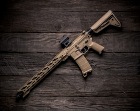 A tan-colored tactical rifle is placed diagonally on a dark wooden surface. The rifle features a sleek design with a detailed handguard, adjustable stock, and a mounted optic. The texture and grain of the wood create a contrasting backdrop for the firearm.