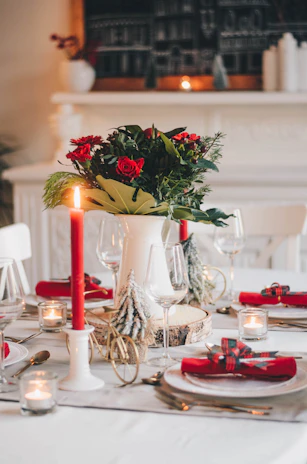 A festive table centerpiece featuring red and green plates, elegant servilletas, and tall velones casting a soft glow.